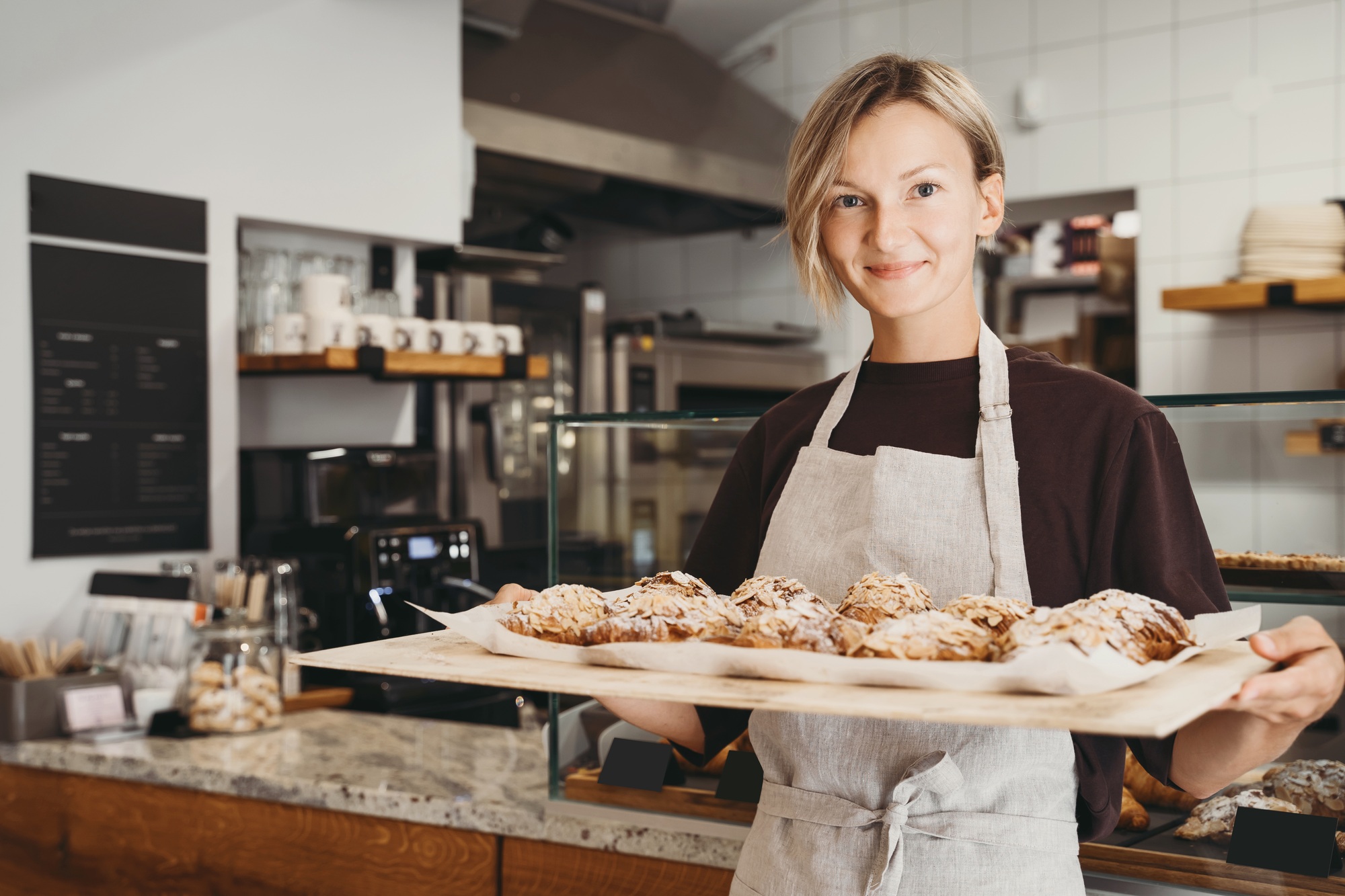 Welcoming female baker holding freshly baked almond croissants in background of bakery shop.